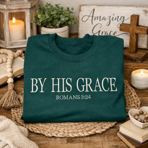 A dark teal folded t-shirt with the text 'BY HIS GRACE ROMANS 3:24' printed in white, displayed on a woven placemat on a rustic wooden table surrounded by candles, books, and a cross.