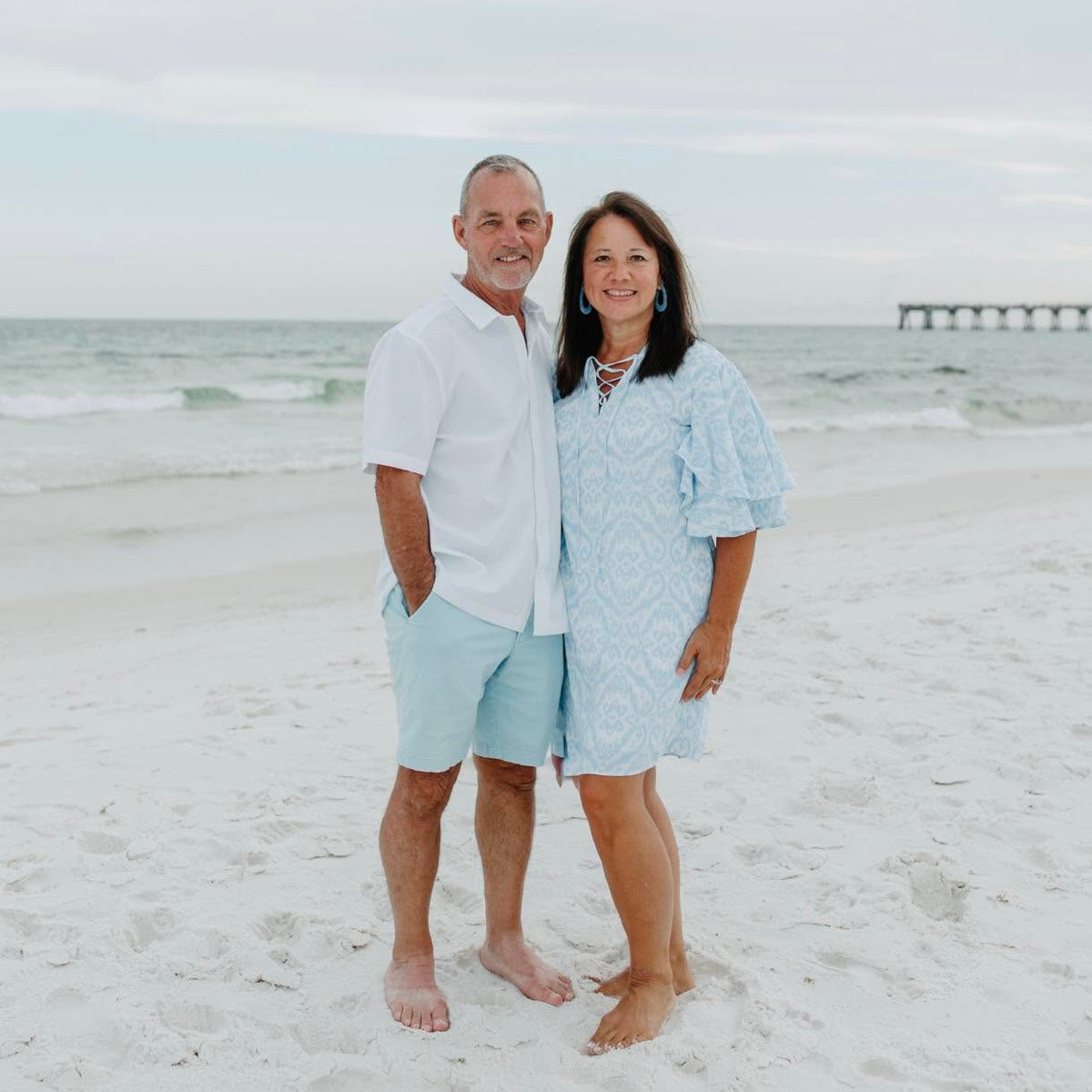 Needles & Knots Owner
A couple standing on a white sand beach with the ocean and a pier in the background. The man wears a white short-sleeve button-up shirt and light blue shorts, and the woman wears a light blue patterned short-sleeve dress.