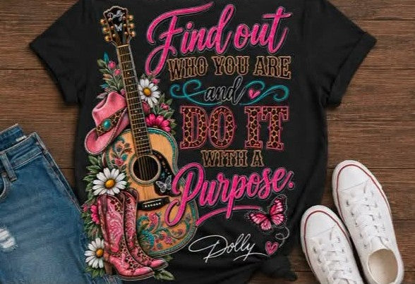 A black graphic t-shirt featuring a western-themed design with a guitar, cowboy boots, flowers, and the text 'Find out WHO YOU ARE and DO IT WITH A Purpose.' by Dolly, styled in a flat lay with blue jeans and white sneakers on a wooden background.