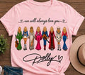 A light pink short-sleeve graphic T-shirt laid flat on a wooden surface, featuring the phrase 'we will always love you' above a row of illustrated women in colorful dresses and 'Dolly' in script below. A tan hat, denim shorts, and a small plant are partially visible around it as styling props.