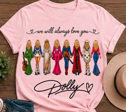A light pink short-sleeve graphic T-shirt laid flat on a wooden surface, featuring the phrase 'we will always love you' above a row of illustrated women in colorful dresses and 'Dolly' in script below. A tan hat, denim shorts, and a small plant are partially visible around it as styling props.