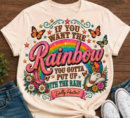 A cream-colored graphic t-shirt featuring a colorful design with the text 'IF YOU WANT THE Rainbow YOU GOTTA PUT UP WITH THE RAIN' and 'Delly Parsons' on a ribbon banner. The shirt is styled in a flat-lay arrangement with blue jeans, white sneakers, and a small plant on a wooden background.