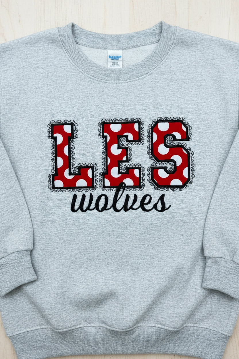 A light heather gray sweatshirt on a wood surface, featuring large decorative letters spelling 'LES' in red with white polka dots and black lace trim, with 'wolves' in black script below.