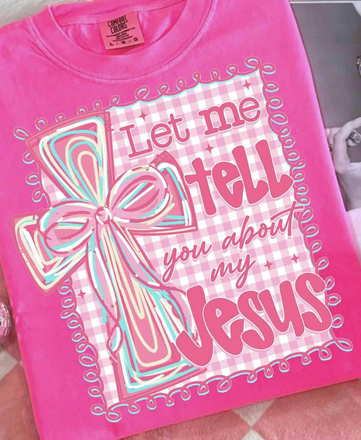 A bright pink short-sleeve t-shirt featuring a graphic design with the text 'Let me tell you about my Jesus' in stylized lettering, alongside a large decorative bow illustration on a pink-and-white gingham background.