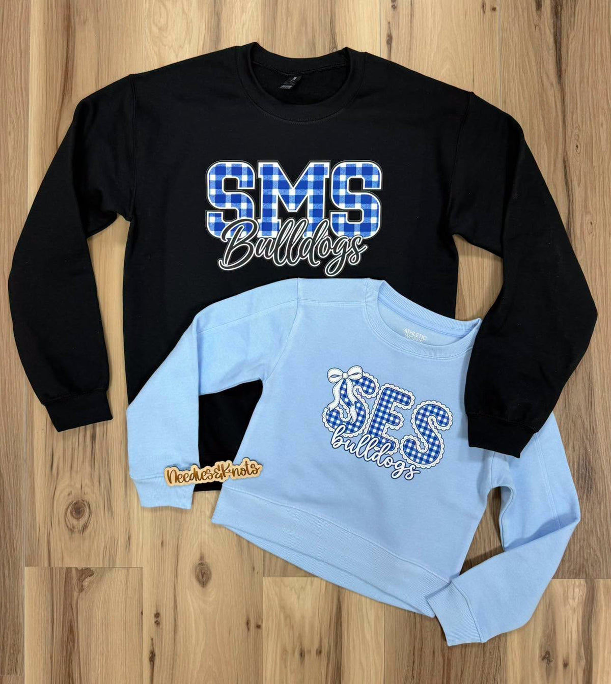 Two long-sleeve sweatshirts laid flat on a wood floor: a black crewneck sweatshirt with a large blue-and-white plaid 'SMS Bulldogs' graphic, and a light blue children's sweatshirt with a smaller matching 'SES Bulldogs' plaid graphic. A small 'TechnoThreads' logo appears near the lower left of the black sweatshirt.
