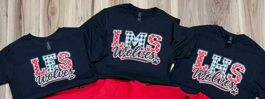A collection of LHS Wolves t-shirts in black and red, featuring various team graphics and logos, arranged on a light wood floor.