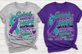 Two graphic T-shirts displayed side by side on a white wood background: a heather gray shirt on the left and a purple shirt on the right, both featuring a teal and purple awareness ribbon with the text 'Suicide Awareness Hope for Today Strength for Tomorrow' and supportive phrases below.