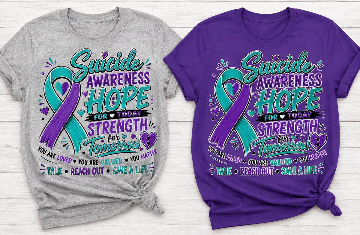 Two graphic T-shirts displayed side by side on a white wood background: a heather gray shirt on the left and a purple shirt on the right, both featuring a teal and purple awareness ribbon with the text 'Suicide Awareness Hope for Today Strength for Tomorrow' and supportive phrases below.