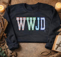 A dark crewneck sweatshirt laid flat on a rustic stone surface, featuring the letters 'WWJD' in a colorful, textured mosaic pattern across the chest. The sweatshirt is styled with candles, an open bible, a cross, and other vintage props.