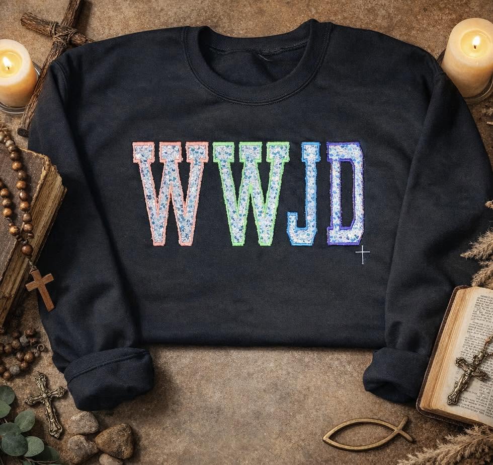 A dark crewneck sweatshirt laid flat on a rustic stone surface, featuring the letters 'WWJD' in a colorful, textured mosaic pattern across the chest. The sweatshirt is styled with candles, an open bible, a cross, and other vintage props.